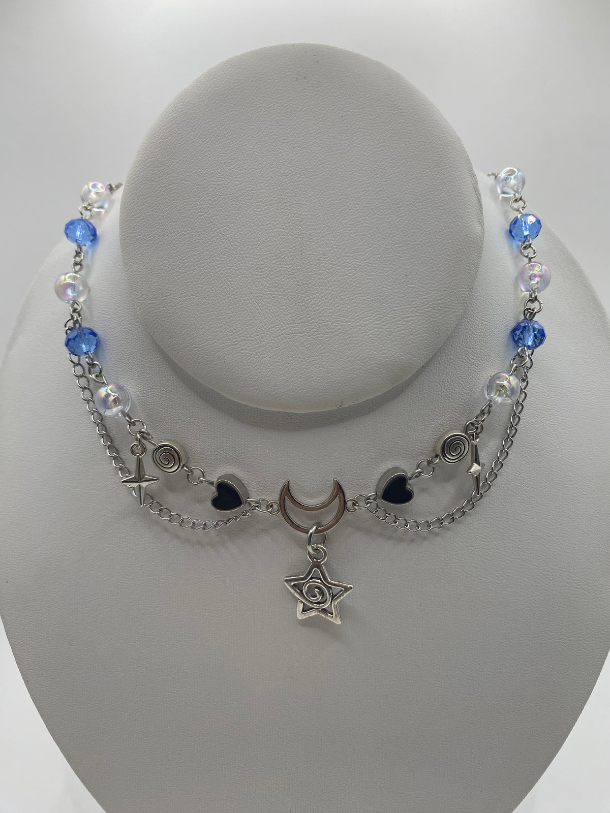 A silver-toned beaded necklace with blue beads, clear iridescent beads, a crescent moon charm, and a swirly star pendant, displayed on a white jewelry bust.