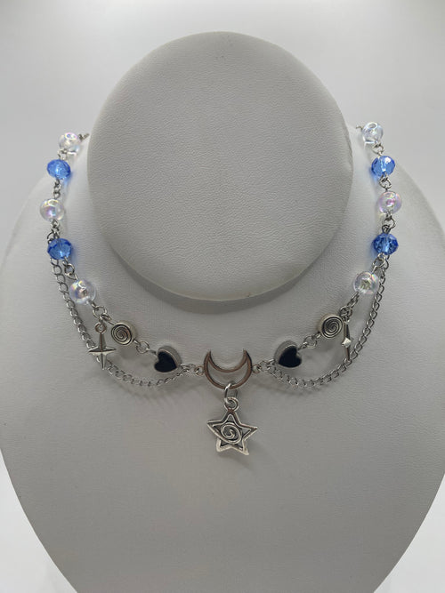 A silver-toned beaded necklace with blue beads, clear iridescent beads, a crescent moon charm, and a swirly star pendant, displayed on a white jewelry bust.