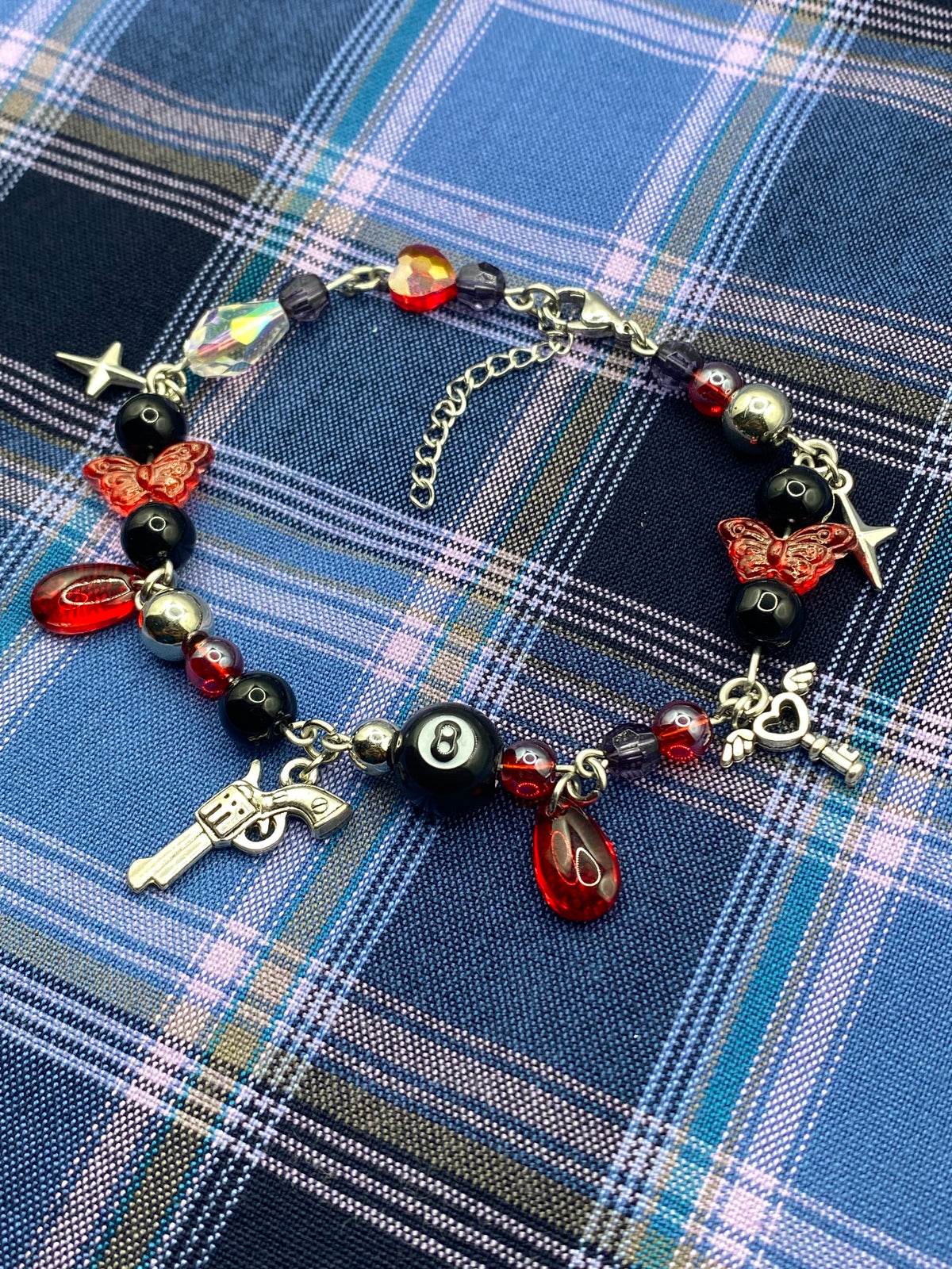 A gothic-style beaded bracelet with red and black beads, skull and butterfly charms, a silver revolver charm, and a black-and-white 8-ball charm, displayed on a blue and white plaid fabric background.