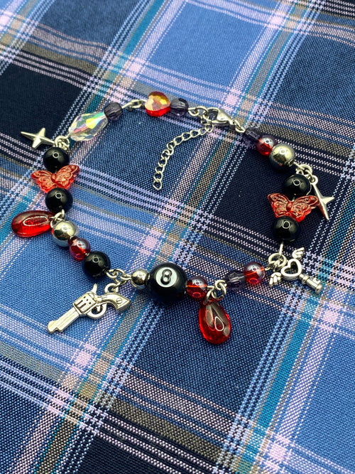 A gothic-style beaded bracelet with red and black beads, skull and butterfly charms, a silver revolver charm, and a black-and-white 8-ball charm, displayed on a blue and white plaid fabric background.