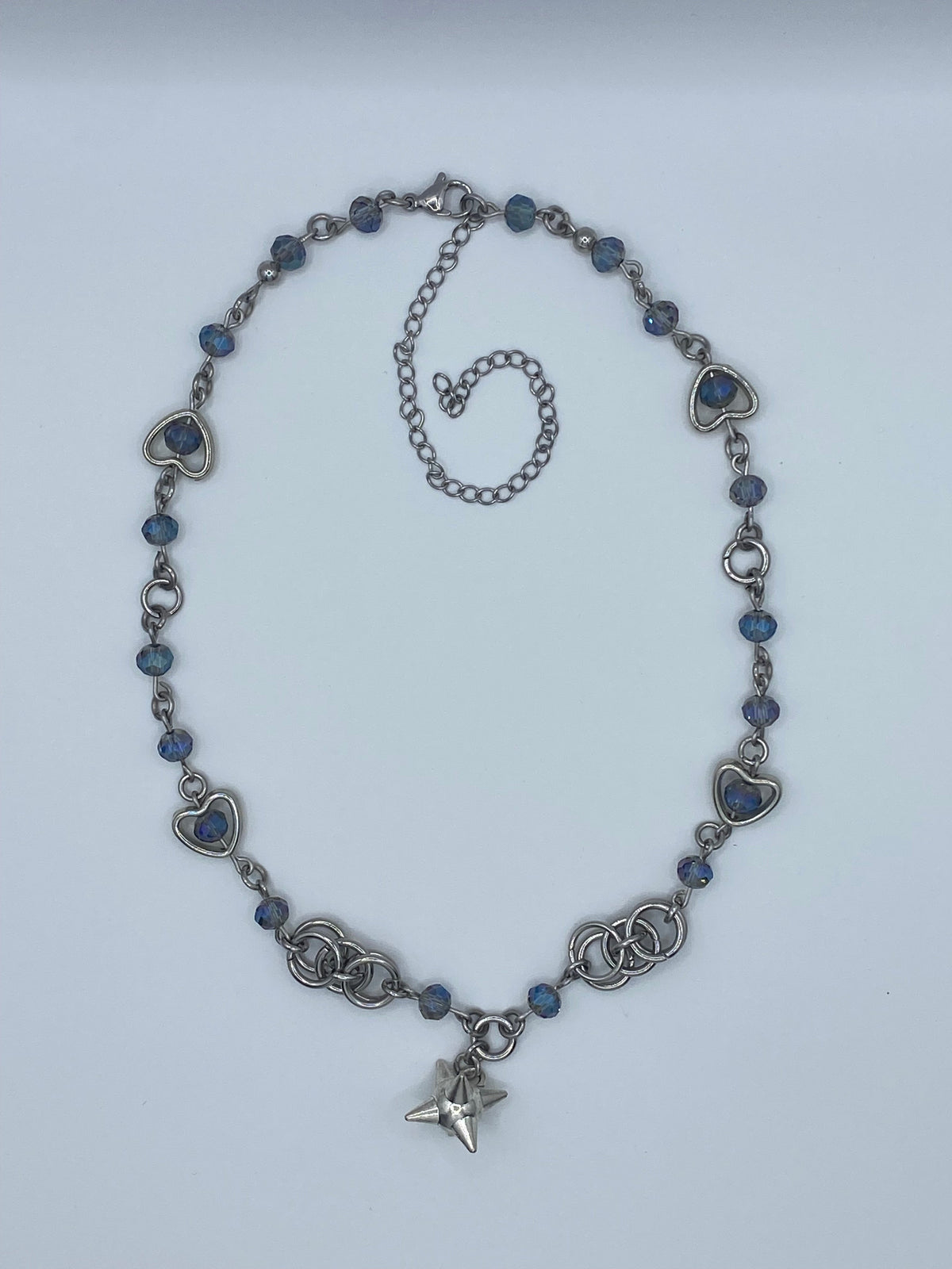 A silver-toned beaded necklace featuring heart-shaped links, faceted blue beads, and a star charm at the clasp, displayed on a light gray background.