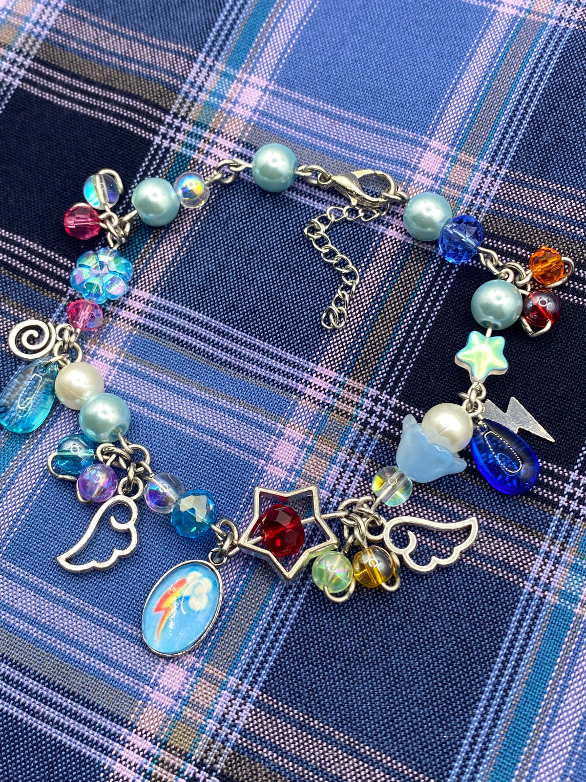 A silver charm bracelet styled on a blue and purple plaid fabric background. The bracelet features various charms including angel wings, a rainbow equestrian oval charm, a red star, a blue cloud, a lightning bolt, and pastel pearls and faceted beads.